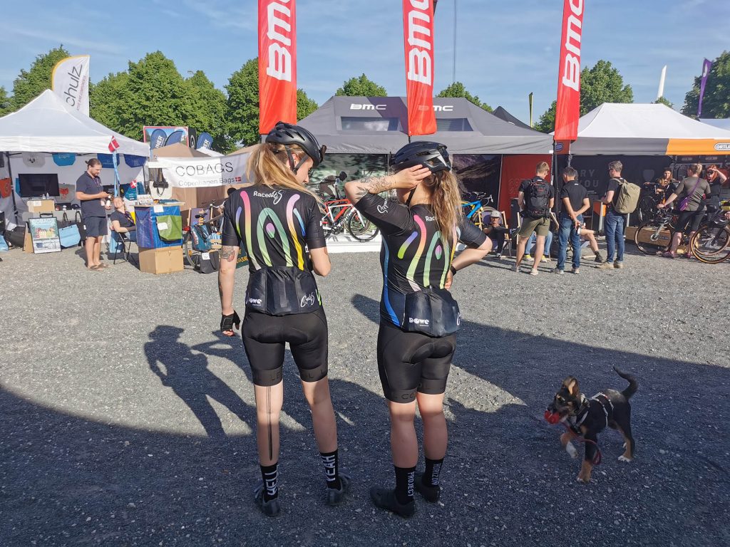Zwei Damen von Team Raceclits in ihren schwarzen Radmonturen von DOWE Sportswear. Sie stehen mit dem Rücken zur Kamera und im Hintergrund sind Zelte und weitere Athleten zu sehen.