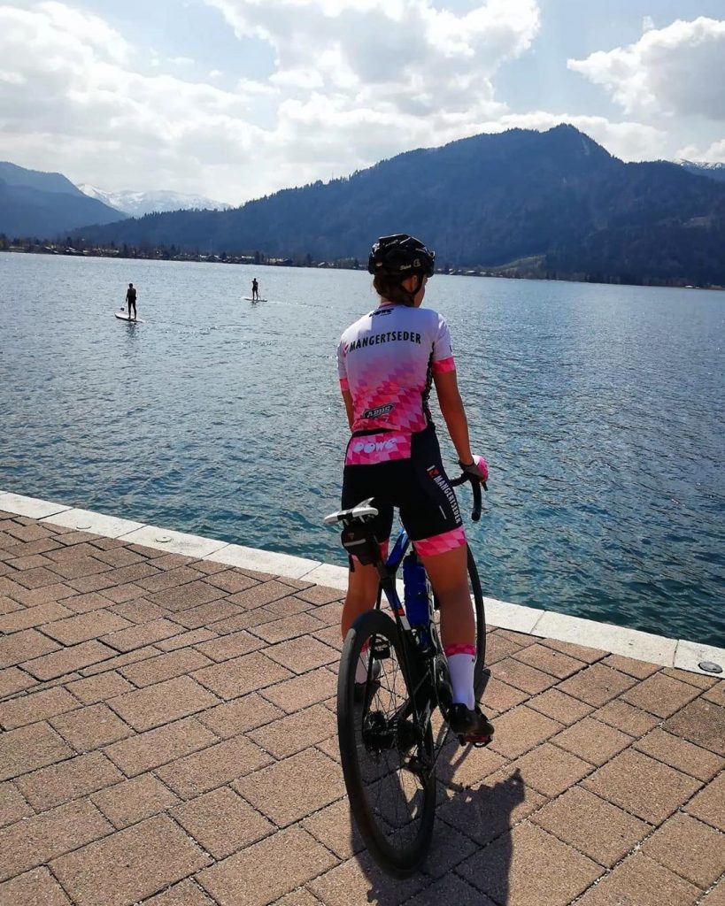 Eine Radsportlerin auf einem Rennrad steht an einem See. Sie trägt ein Trikot von Team Mangertseder, produziert von DOWE Sportswear. Das Trikot ist pink.