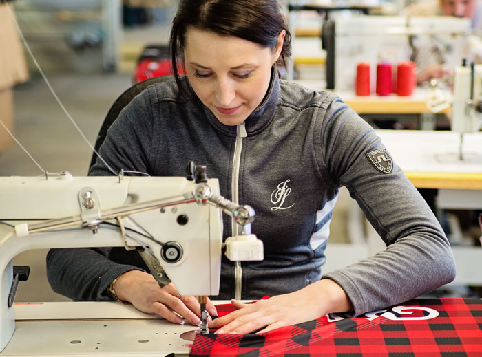 Eine Näherin sitzt in einer Näherei und näht ein rotes Teamwear-Kleidungsstück von DOWE Sportswear. Sie blickt konzentriert und zufrieden auf ihre Arbeit.
