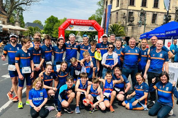 dowe-sportswear-S30021+S30022-IMG-20230509-WA0002-bb Eine große Gruppe von jungen und mittelalten Athlet*innen in blauen Funktionsshirts von DOWE. Sie posieren sitzend und stehend vor einer Zielmarkierung, vermutlich bei einem Stadtlauf