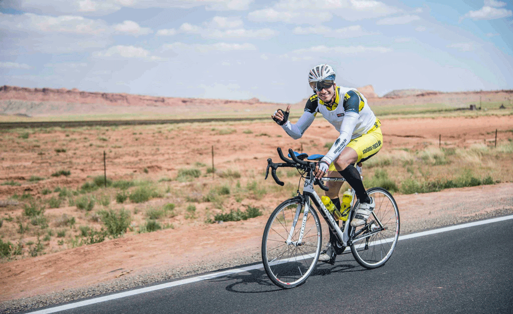 Ultra-Cyclist Stefan Schlegel fährt auf einem Rennrad über eine Straße in der Wüste. Er trägt gelb-weiße Teamwear von DOWE und hält seinen rechten Daumen hoch.