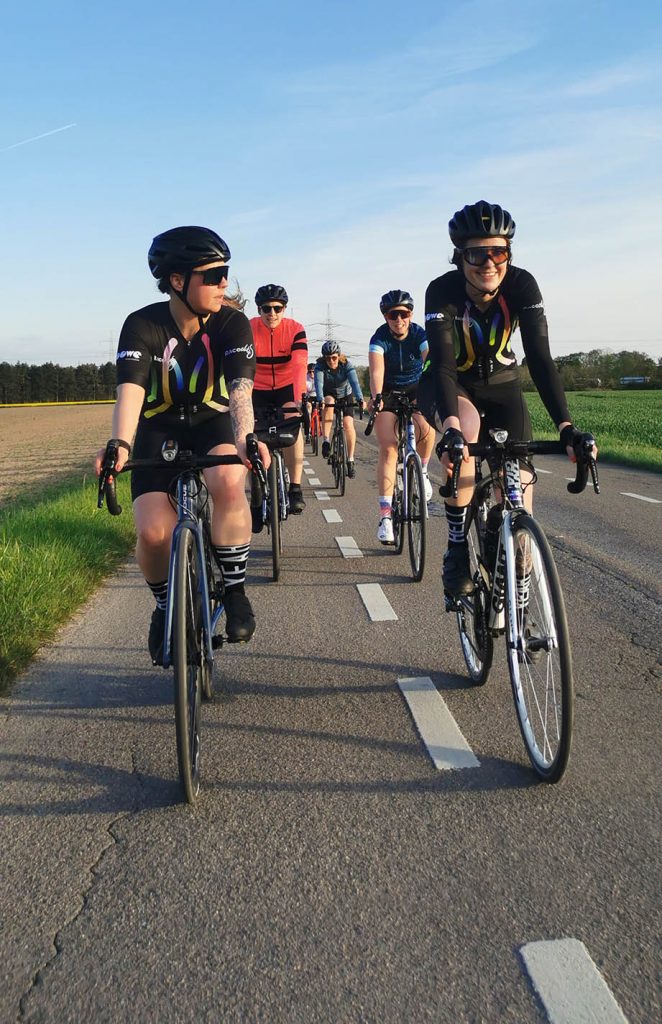 Das Damen-Team "Raceclits" in Trikots von DOWE Sportswear auf ihren Rennrädern - Sie fahren auf einer geteerten Straße richtung Kamera.