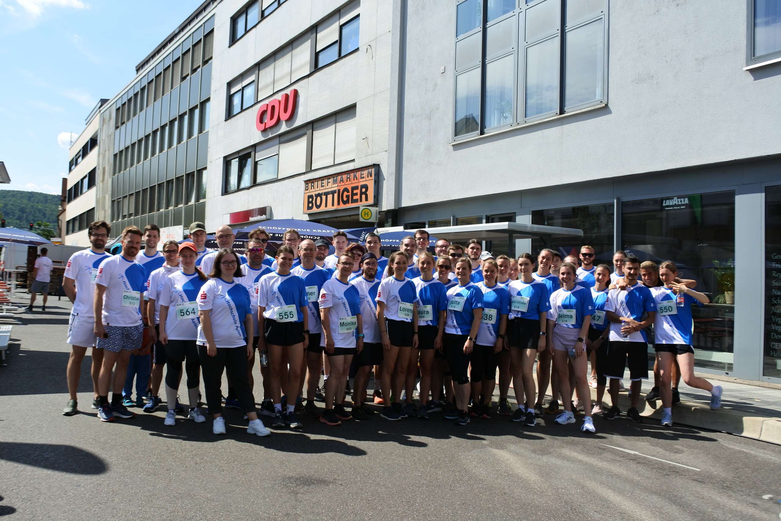 Ca 40 Athlet*innen mit blau-weißen Laufshirts von DOWE Sportswear stehen posierend auf einem Platz. Es ist sonnig.
