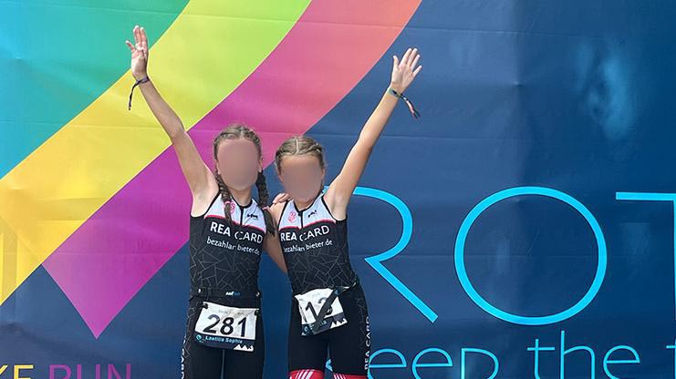 Zwei junge Athletinnen in DOWE Sportanzügen heben jubelnd die Hände vor einer bunten Sponsoren-Wand