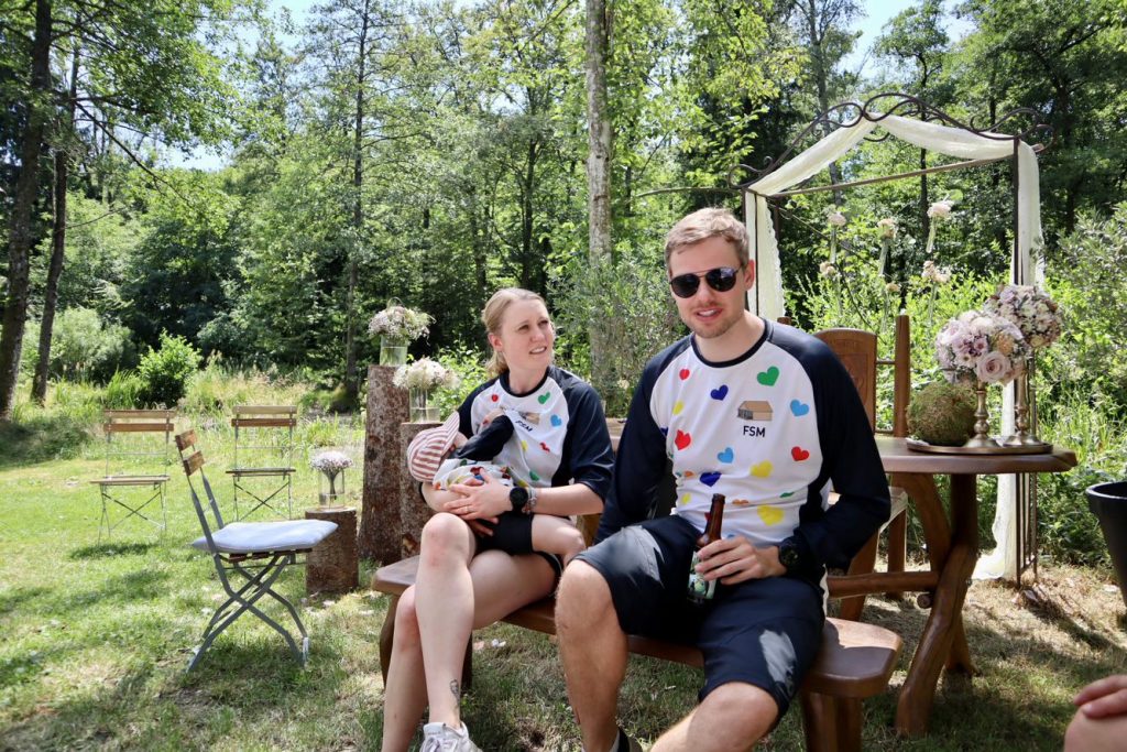 Ein Brautpaar sitzt, gekleidet in Bike-Jerseys von DOWE in einem Garten. Sie haben ein Kleinkind bei sich; er trägt Sonnenbrille und trinkt Bier.