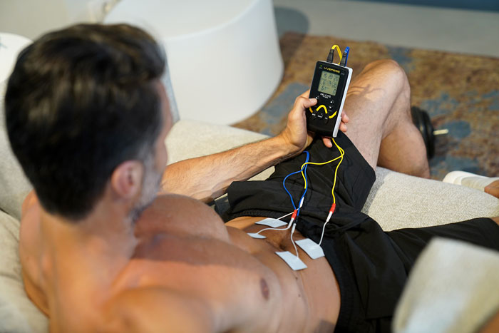 Ein athletischer Mann mit nacktem Oberkörper bedient ein Gerät "Mio Care Fitness" in seiner Hand. Kabel davon sind an seinem Bauch befestigt.