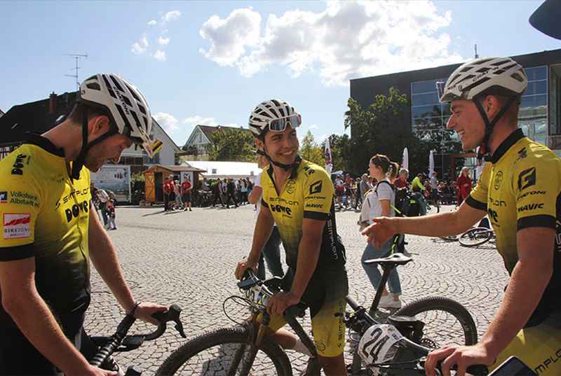 Team DOWE Simplon - Drei Radler im Trialog auf einem öffentlichen Platz mit Pflastersteinen
